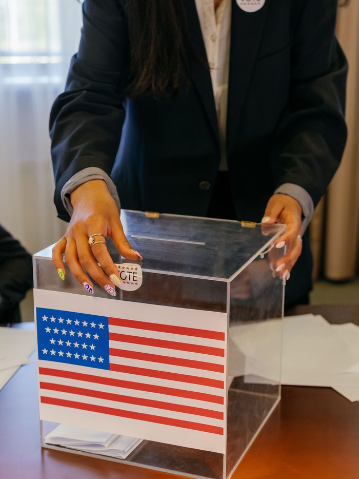 An election worker steadying a clear ballot box marked with a United States flag
