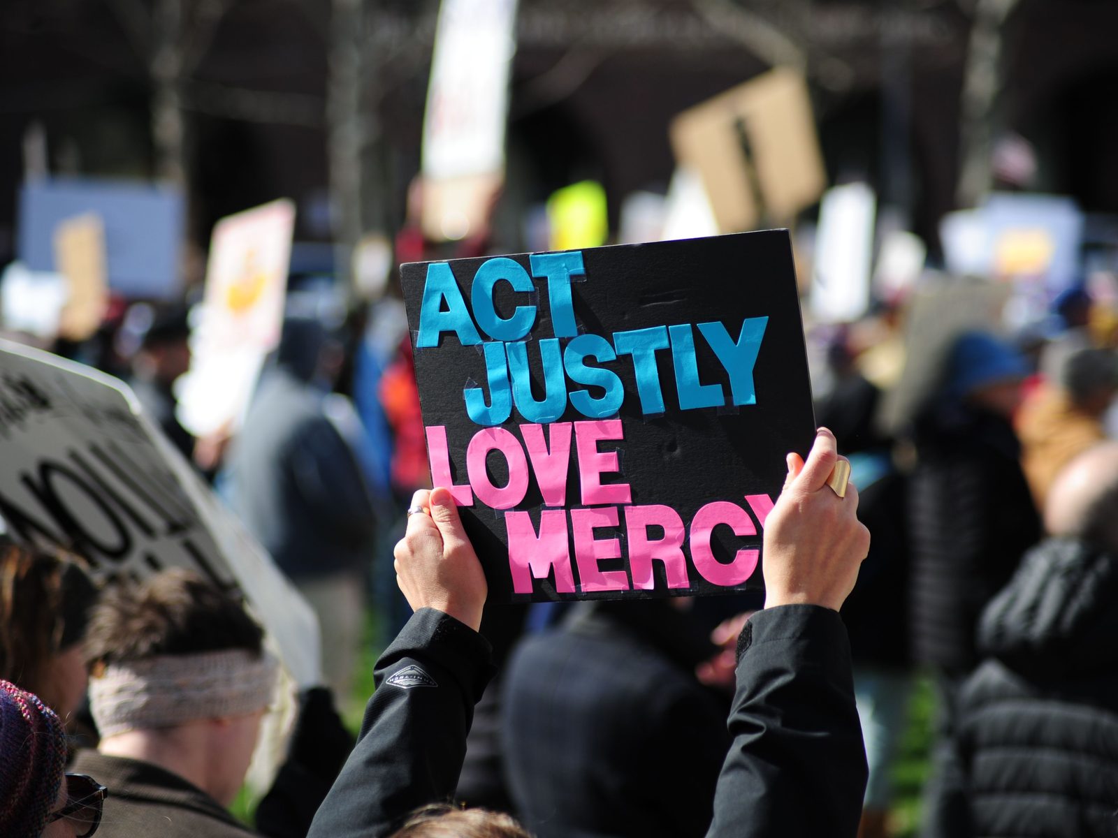 A demonstrator holds a hand-lettered sign reading Act Justly, Love Mercy above a crowd at a public rally