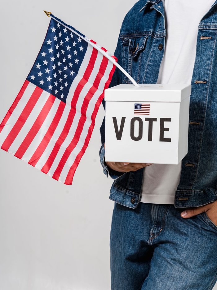 A person holding a ballot box marked VOTE alongside a United States flag