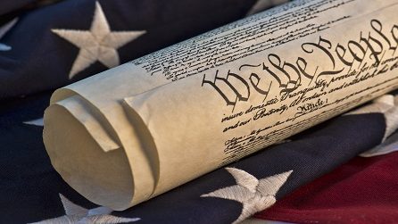 A rolled copy of the United States Constitution resting on a flag, the words We the People visible